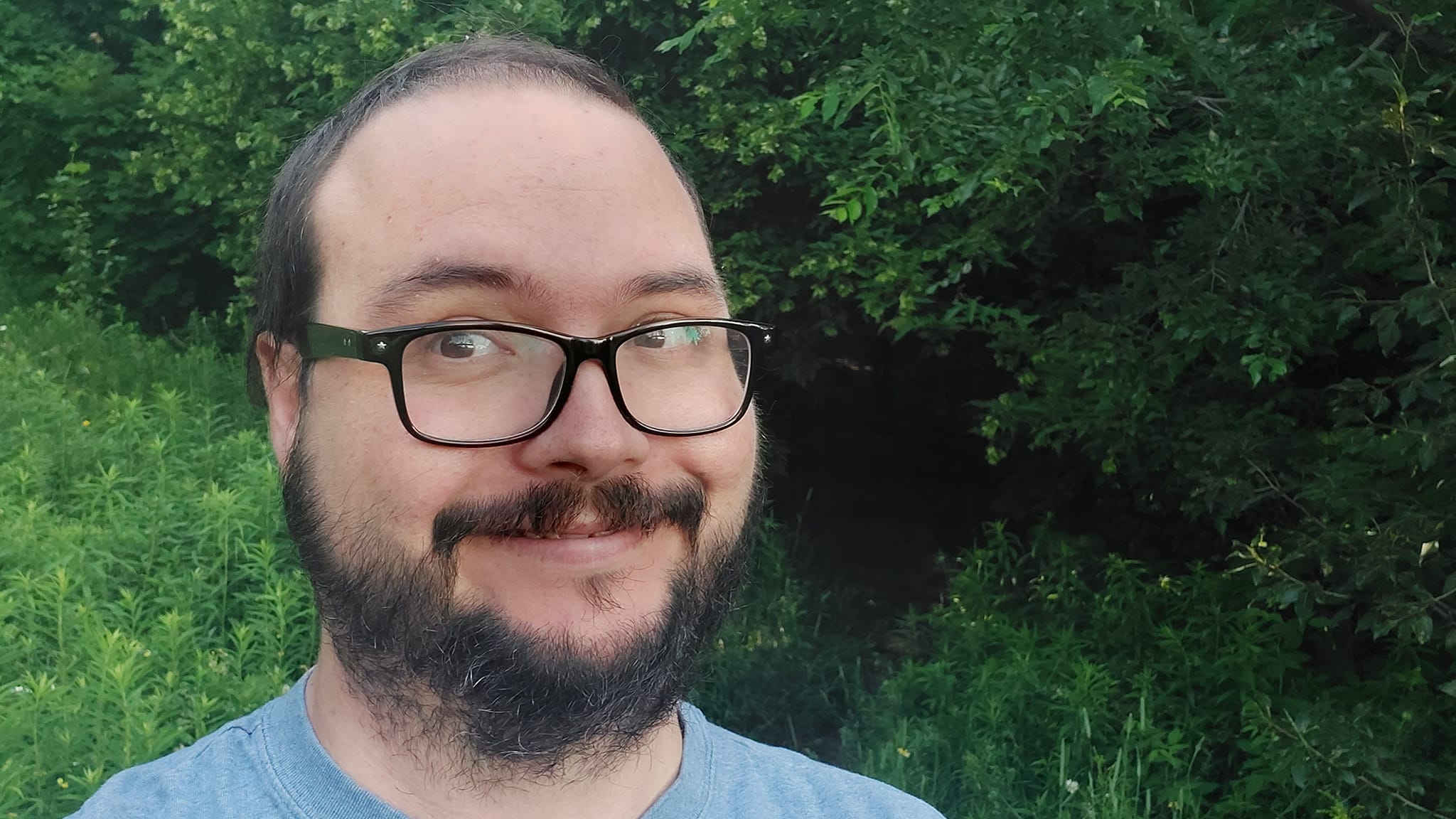 A man is dressed in a t-shirt standing in a field in front of a path through the trees